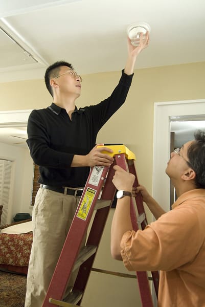 man holding ladder for another man installing a smoke alarm